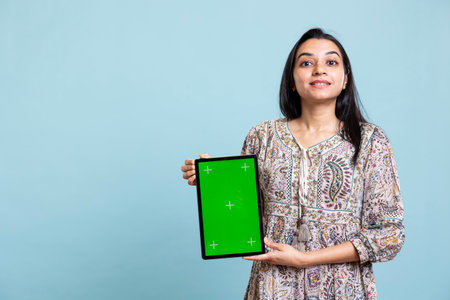 Indian woman showing isolated copy space display in front of the camera, creating an advertisement for a commercial against blue background. Young joyful adult holding device in studio.の写真素材
