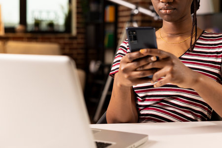 Close up of african american businesswoman using smartphone to respond to company messages. Black female entrepreneur texting on mobile device, managing communication while working remotely.の写真素材