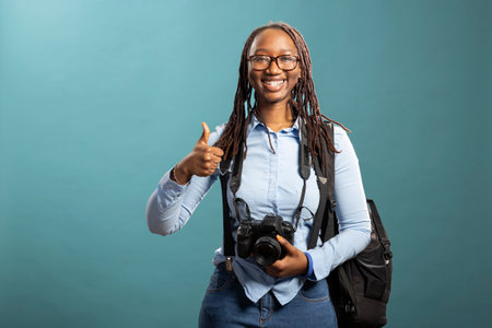 Energetic female photographer in studio with camera around her neck, giving thumbs up, excited for an upcoming photoshoot. African american woman doing an approval gesture against isolated background.の写真素材
