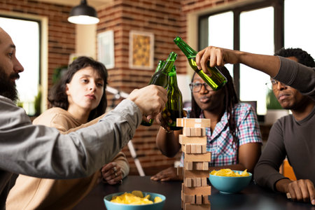 Group of diverse friends clinking beer bottles, seated around table with snacks and games. Caucasian and african american people enjoying an indoor hangout in warm, relaxed atmosphere.の写真素材