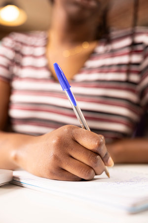 Selective focus on hand of female employee using pen to write down marketing strategy ideas on notepad. Close up of self employed black woman planning work schedule at home office desk.の写真素材