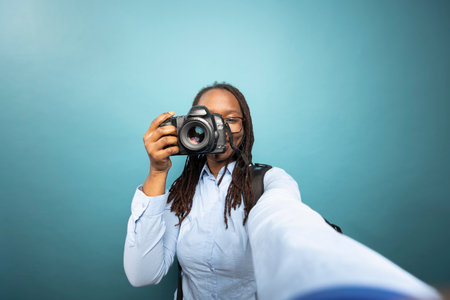 Black female photographer records herself, smiling toward device while holding DSLR close to her...