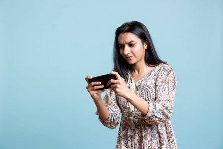 Ecstatic woman playing mobile video games on her smartphone, playing to defeat foes and clenching her hands. Indian female gamer enjoying leisure activity during her free time in the studio.の写真素材