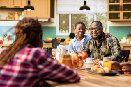 African American family sharing pastry and drinks for breakfast at home, parents serve scrambled eggs, toast and fruit. Cozy kitchen moment captures warmth, relaxation and joyful togetherness.の写真素材