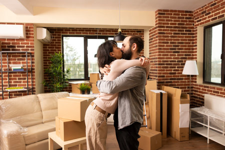 Caucasian couple kisses and embraces in living room after purchasing apartment, excited to move in together. Carefree husband and wife share tender moment surrounded by cardboard boxes in new home.の写真素材