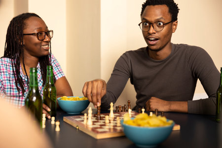 African american man with glasses moving a chess piece with shocked facial expression. Young black male individual looking surprised during casual game with friends, snacks and drink nearby on table.の写真素材