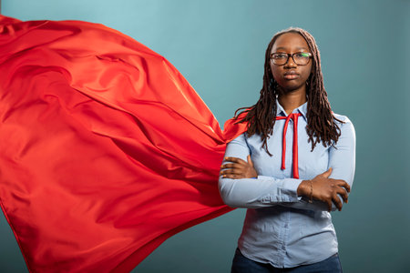 Confident african american woman stands with crossed arms and red cape flowing, striking a power stance in studio. She radiates strength, style and empowerment on blue background.の写真素材