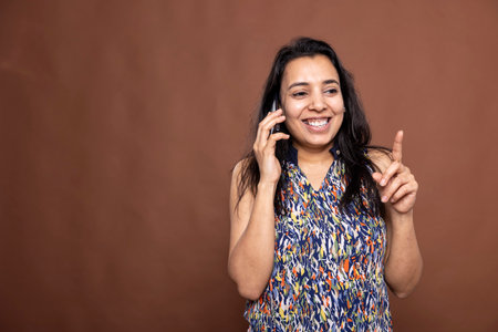 Smiling indian woman holding cellphone to her ear and making hand gestures during the call. Beautiful happy female individual enjoying cheerful conversation with family or friends.の写真素材