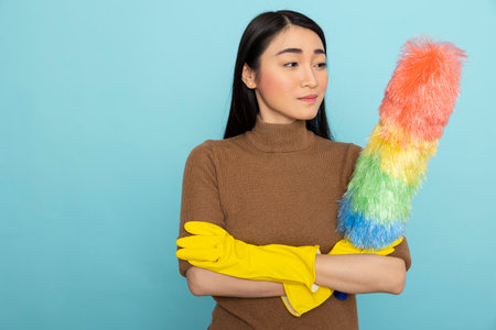 Pretty female janitor wearing yellow rubber gloves and standing with feather duster, prepared for cleaning services. Portrait of asian housekeeper posing with arms crossed, confident in completing chores.の写真素材