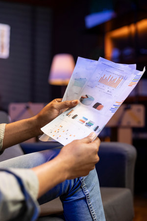 Young adult sitting on the sofa and reading research papers with analytics, multitasking with files and documents at home. Man diving into professional or personal topics of improvement.の写真素材