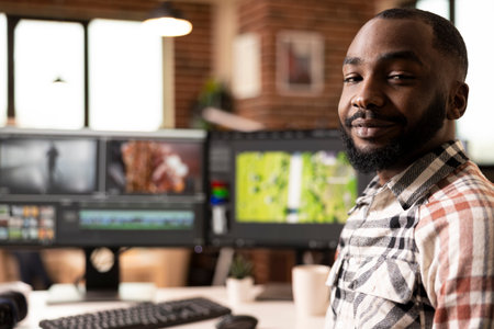 African american male filmmaker smiling confidently at brick wall home studio. Creative black man seated at dual monitors with color grading software, ready to remotely produce cinematic videos.の写真素材