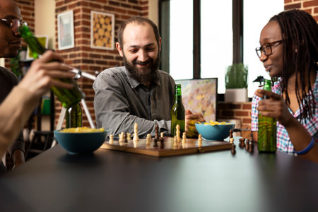 Bearded man looking at chessboard with a smile as his african american friend sit nearby holding a drink. Casual moment among multiethnic individuals enjoying indoor game session with snacks.の写真素材
