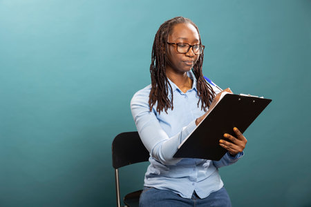 African american woman holds clipboard and pen, seated and writing on paper against isolated background. Young black female individual takes notes, documenting information on notepad in studio.の写真素材