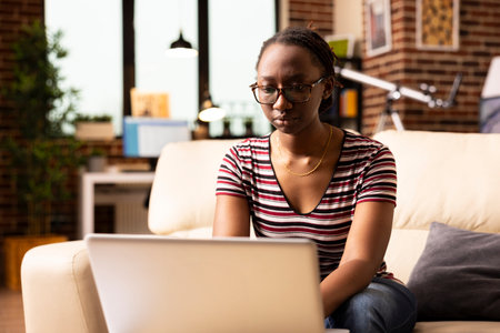 African american freelancer working on clientele business project presentations, focused on providing good online services. Black woman seated on couch, answering emails on laptop.の写真素材