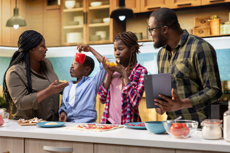 Black adorable parents and kids following online cooking recipe for pizza day, enjoying fun weekend activity making homemade pizza together in the kitchen. Happiness and teamwork for bonding.の写真素材