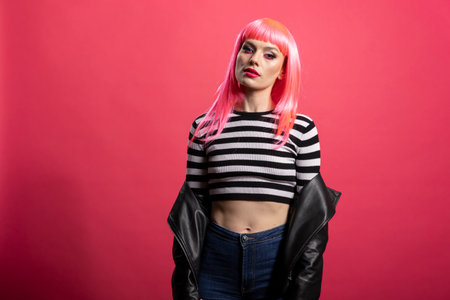 Gorgeous white woman with vibrant hairstyle and striped crop top stands poised and looks at camera. Beautiful young lady with black jacket, expresses calm confidence in pink background studio.の写真素材