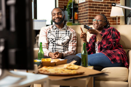 Smiling black man sitting on sofa beside girlfriend with remote, watching movie at home. Young couple relaxing with digital entertainment and takeout food during weekend in brick wall apartment.の写真素材