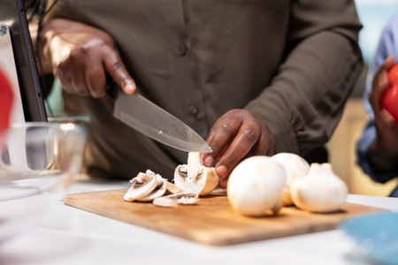 Close up of african american mother cutting mushrooms in the kitchen, preparing the ingredients for homemade pizza and teaching children the old family recipe. Fun parenting and bonding.の写真素材