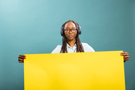 Young black woman with wireless headphones, standing and holding yellow banner. African american female individual listens to music, grasping blank placard and looking at camera. Ideal for ads.の写真素材