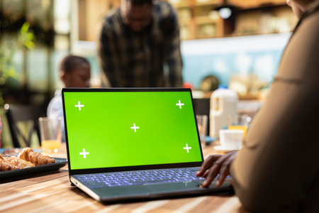 Green screen laptop next to parents and kids sharing breakfast in the kitchen, serving eggs, croissants and orange juice. Bright and cozy domestic moment celebrates family bonding together.の写真素材