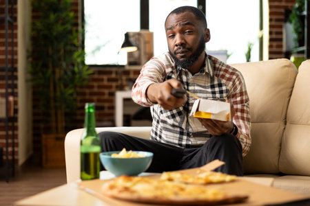 Young african american man seated on couch squinting at TV while flipping channels. Black guy holds remote control and takeaway food box in relaxed living room, enjoying casual meal at home.の写真素材