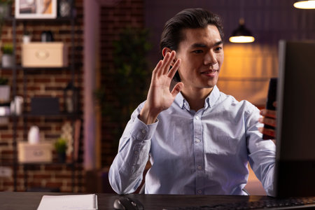 Young businessman waves at smartphone, joining video call from home. Asian male entrepreneur communicates virtually with colleagues, enjoying remote work and staying connected in cozy apartment.の写真素材