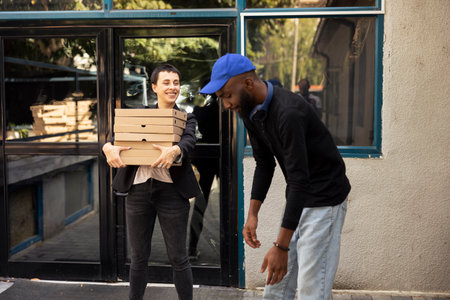 Black delivery worker passes stack of hot pizzas to client at the doorstep, restaurant takeaway service. Fast food large cardboard boxes order delivered directly to a woman customer.の写真素材