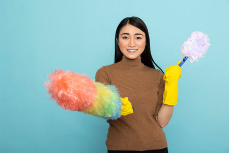 Happy asian woman holding two brushes, ready to start morning dusting routine. Confident female janitor with rubber gloves, smiling brightly and grasping cleaning tools against isolated background.の写真素材