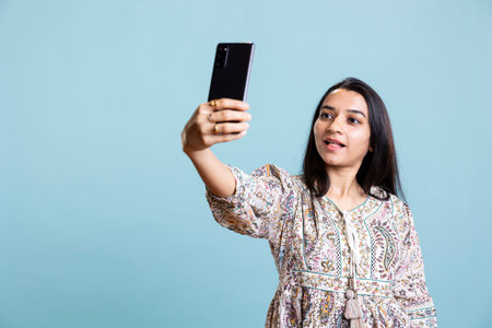 Smiling confident person takes pictures on her mobile phone app, feeling pretty and taking photos against blue background. Young indian woman showing joy and a positive attitude.の写真素材