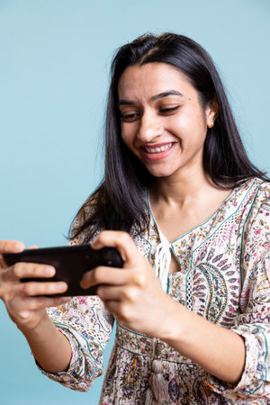 Confident happy player smiling and paying attention on the video gaming competition, e sport tournament on her mobile phone. Young indian woman enjoys interactive game in studio.の写真素材