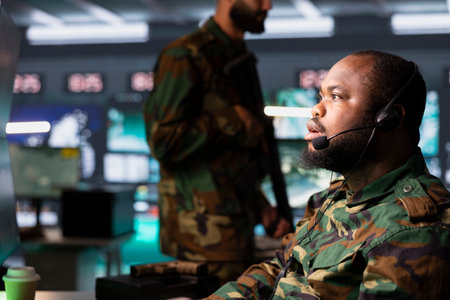Army dispatcher looking at surveillance transmission on screens, relaying data to troops using headset mic. African American military operative transmitting reconnaissance infoの写真素材