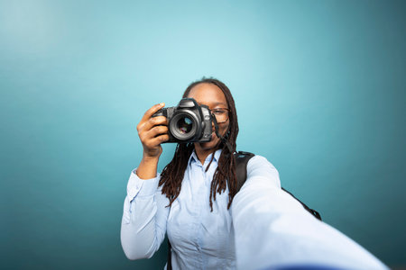POV of Black female photographer recording herself with a DSLR. African American woman in blue shirt, smiling while documenting her photography journey in a studio with confidence.の写真素材