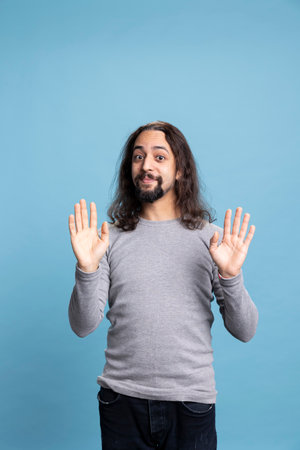Silly young adult making goofy faces and acting innocent against blue background, joking around and...
