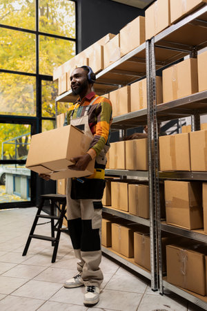 African american worker carries cardboard boxes with tunes on headphones, wearing coveralls and preparing goods for shipment. Arranging parcels on shelving units for easy distribution.の写真素材
