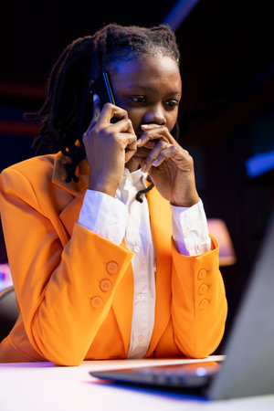 Woman seated at home office desk using laptop, answering phone call while checking emails displayed on screen. Remote worker in telephone call with friend while reading messages in inboxの写真素材