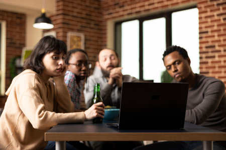 Group of multiethnic friends gathered around table, focused on laptop screen during indoor hangout. Caucasian and african american people enjoying movie together, sharing relaxed moment with drinks.の写真素材