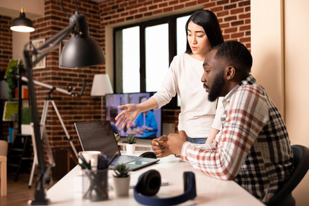Supportive girlfriend discussing software on laptop with focused male developer at home office desk. Multiethnic couple working remotely together and brainstorming on app development for tech company.の写真素材