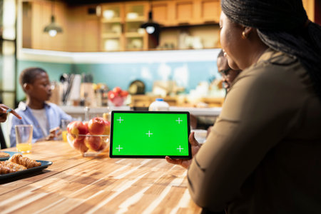 Green screen held by mother with her children sharing breakfast in home kitchen, serving croissants....