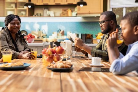 African american dad reading daily newspaper at the kitchen table, enjoying morning routine during breakfast with his little family. Young dad feeling happy chatting with his children and wife, eat together.の写真素材