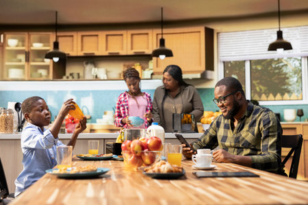 Father working on his smartphone and serving breakfast with his cute family, gathered in the kitchen to enjoy the simple comfort of morning togetherness. Happy start to the day with sunlight.の写真素材