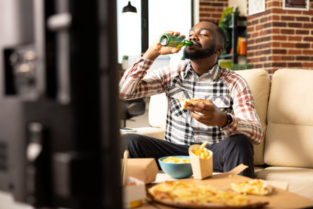 Relaxed weekend of black man sipping beer and eating slice of pizza in cozy brick wall apartment. African american guy watching movies and enjoying takeaway food in leisure time at home.の写真素材