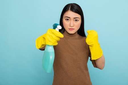 Female cleaner with rubber gloves stands in fight pose with spray bottle, expressing anger and...