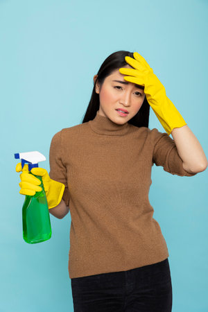 Exhausted asian housekeeper wearing yellow gloves rubs her head while holding green spray bottle. Overworked female cleaner with sad expression, experiencing headache.の写真素材