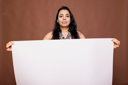 Beautiful female model holding white empty poster, ideal for promotional content in brown studio environment. Pretty woman grasps blank placard in front of her, presenting blank copy space for ads.の写真素材