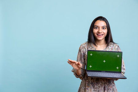 Cheerful indian woman presenting the laptop with isolated mockup screen, showing the display for a marketing commercial in front of the camera. Young adult creating an advertisement.の写真素材