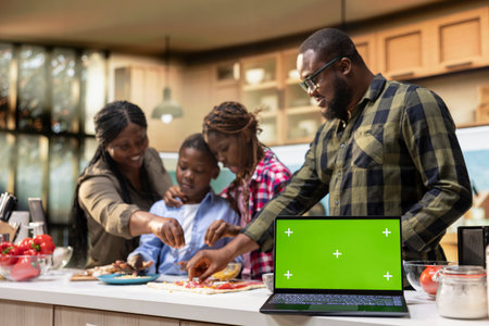 Green screen laptop with parents and children gathered in the kitchen, preparing homemade pizza with fresh ingredients and laughter. Cozy moment celebrates teamwork and delicious food.の写真素材