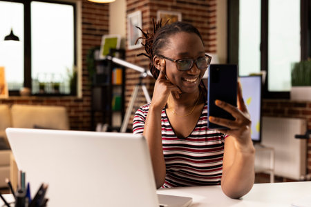 Smiling african american businesswoman seated at desk, having pleasant conversation on mobile device. Remote female employee taking break while using smartphone for video call in apartment.の写真素材