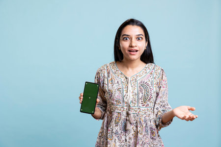 Positive happy girl presenting chroma key isolated screen on device, doing promotion for a commercial against studio background. Upbeat indian person used for advertising.の写真素材