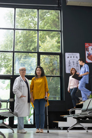 Doctor supporting woman using crutches during rehabilitation session in medical facility hallway. Medical staff offers a helping hand for healing process, improving balance and mobility.の写真素材