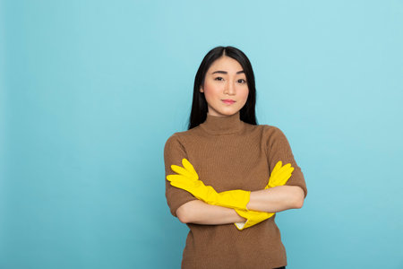 Asian woman in yellow rubber gloves posing with arms crossed, confidently prepared to inspect cleaning results. Female housekeeper stands against blue background, looking at camera.の写真素材
